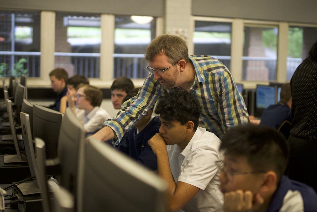 Students sitting at computer screens in a classroom with a man pointing at one of the screens