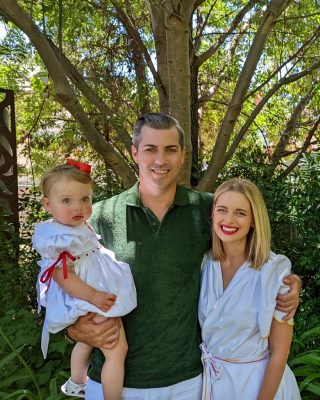 Image of a family in front of a tree. A man in a green shirt is holding a toddler and has his arm around a lady in a white dress. 