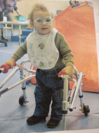 A young baby wears glasses and stands, holding a metal frame to support his walking.