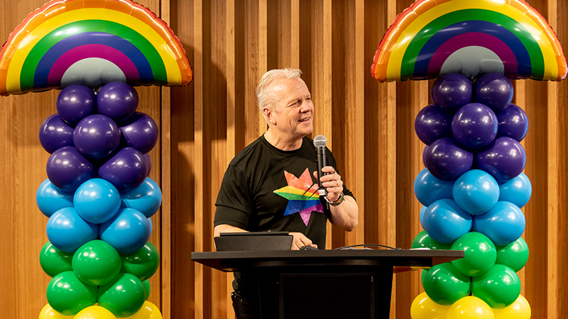 Drew Bradford speaking at a NAB pride event, rainbow balloons behind him