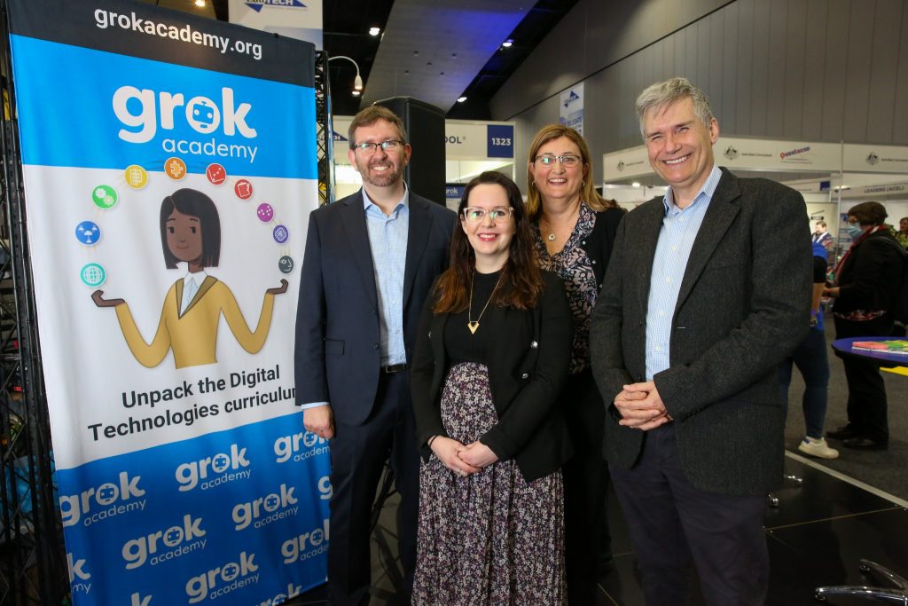 Two women and two men standing, smiling, next to a banner displaying the name 'Grok Academy'