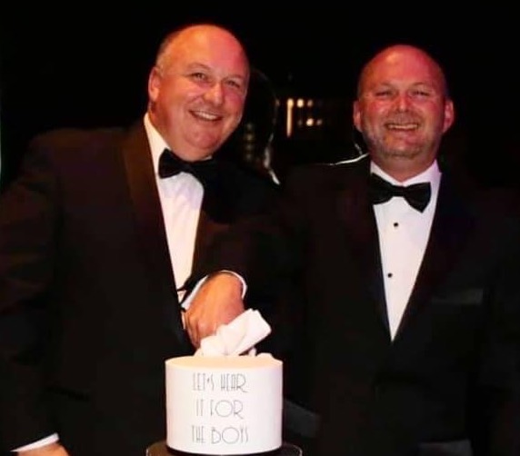 Two men in tuxedos smile at the camera. Their wedding cake is on a table in front of them.