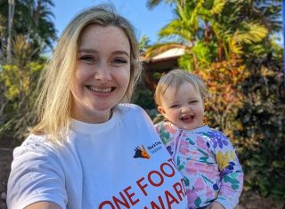 A woman holding her toddler both smile at the camera. The woman's t-shirt has 'One Foot Forward the walk for mental health' printed on the front.