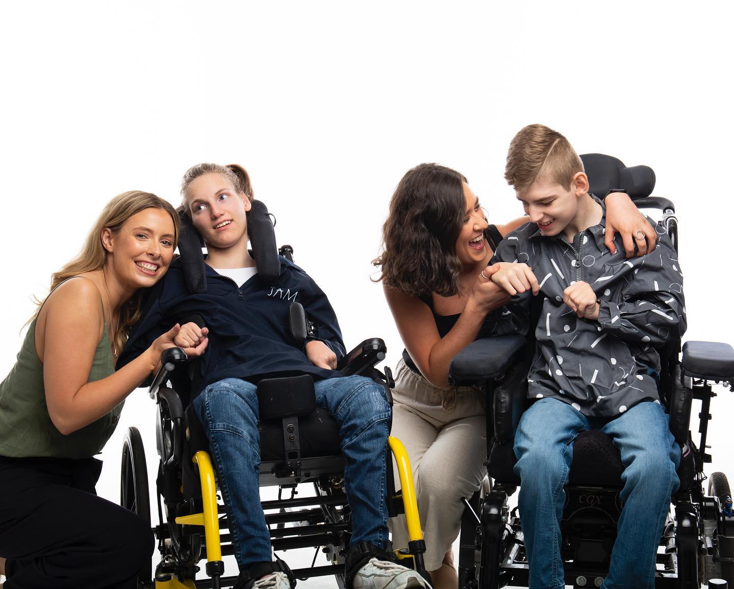 Two women hold hands of a boy and a girl in mobility chairs.