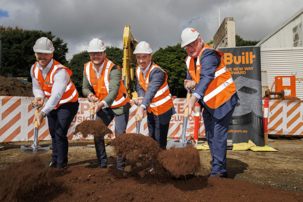 Four men standing side-by-side at a construction site shovelling soil