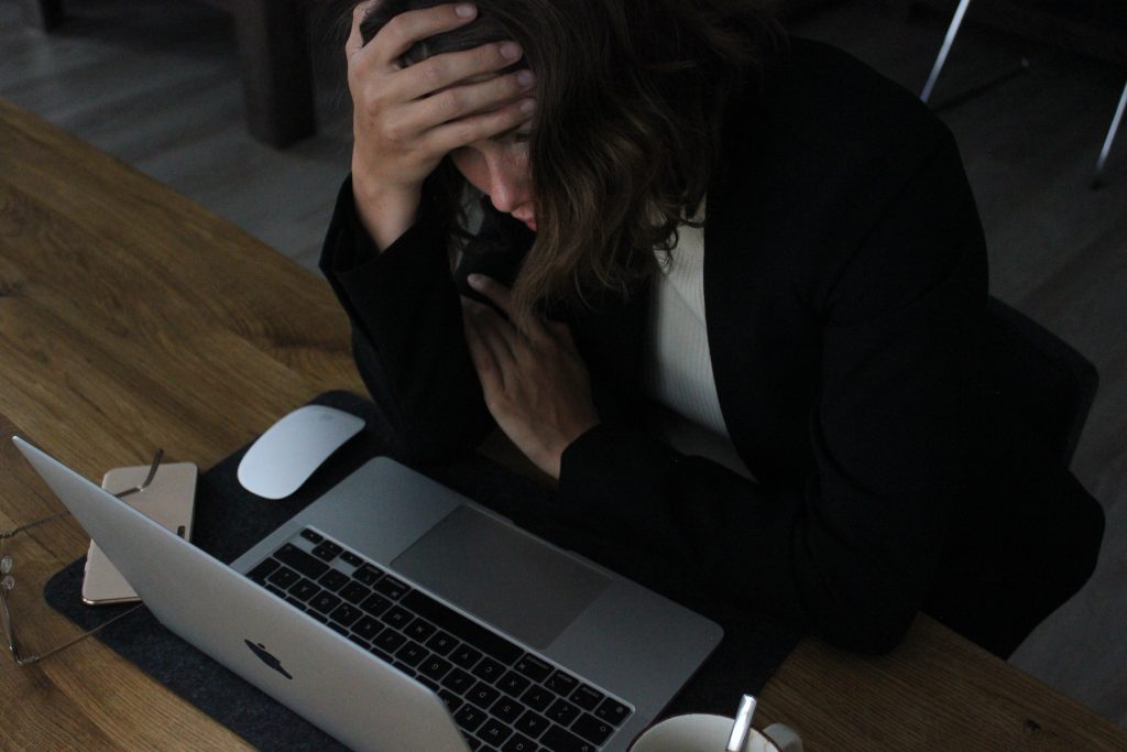 Image of a woman sitting at a table with a computer open and her hand on her forehead