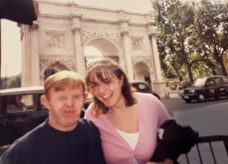 A man and a woman smile in front of a bridge, with cars going past behind them.