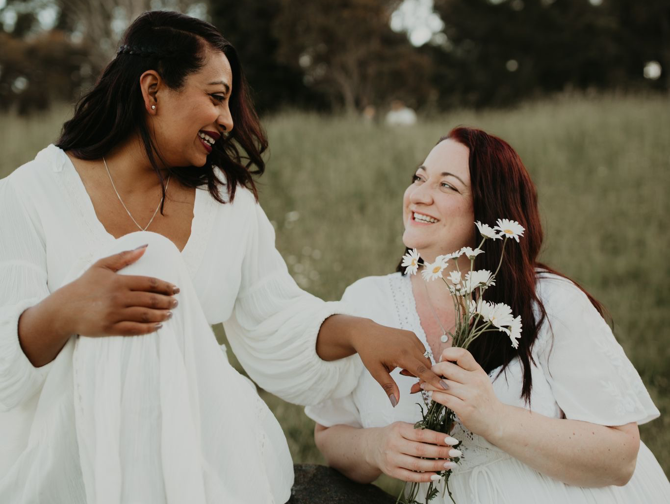 Two women in wedding dresses in a field, holding flowers