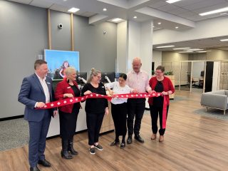 Six people standing side-by-side holding and cutting a ribbon inside a bank branch
