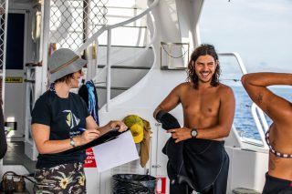 A young man on a boat in a wetsuit, alongside a your woman with a clipboard