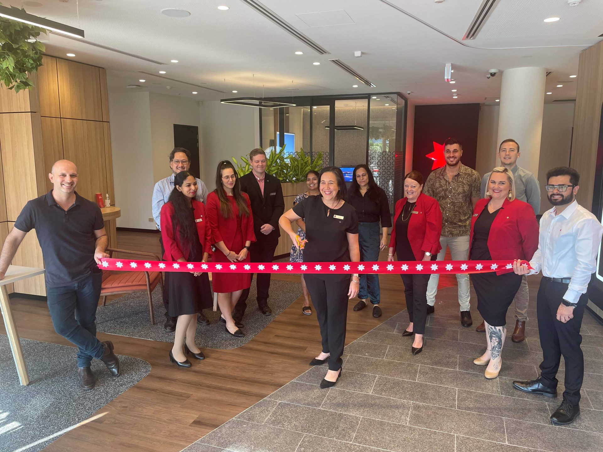A team of 13 people standing behind a red ribbon as the branch manager cuts the ribbon with scissors