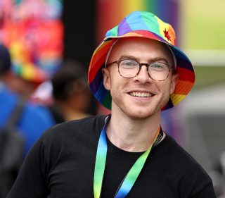 A man smiles at the camera. He is wearing a rainbow hat and lanyard, glasses and a black t-shirt.