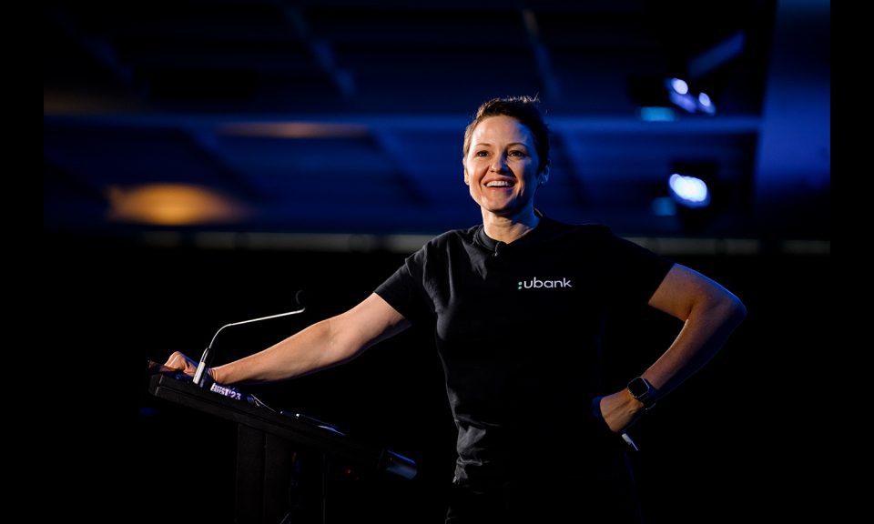 A lady stands at a podium wearing a black t-shirt. It is dark in the background with faint purple light.