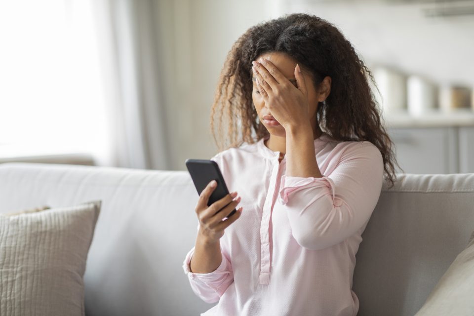 woman on a couch holding a phone in one hand and with her other hand raised to her forehead