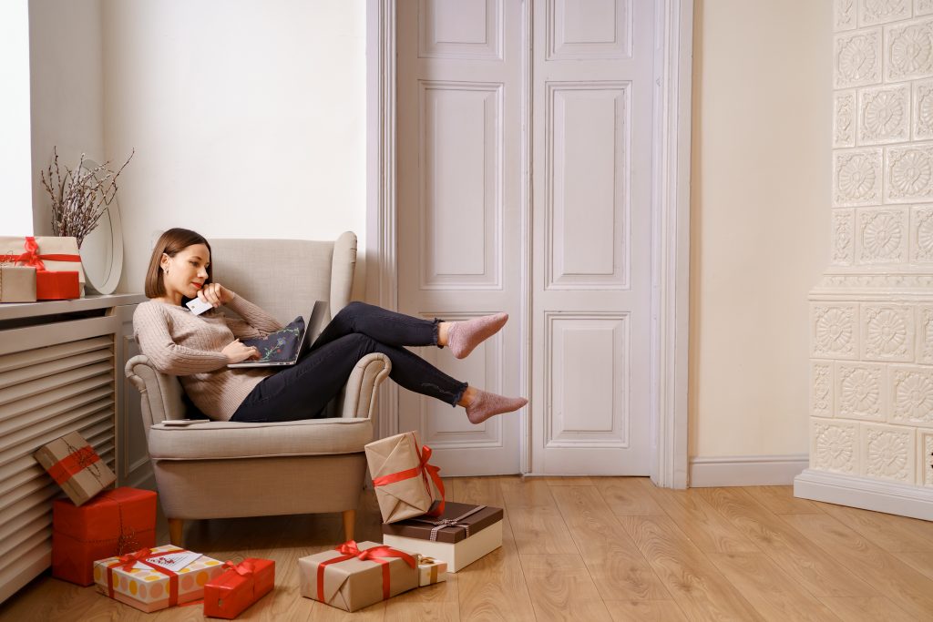 woman on chair with credit / debit card in hand and computer on lap. she is surrounded by presents.