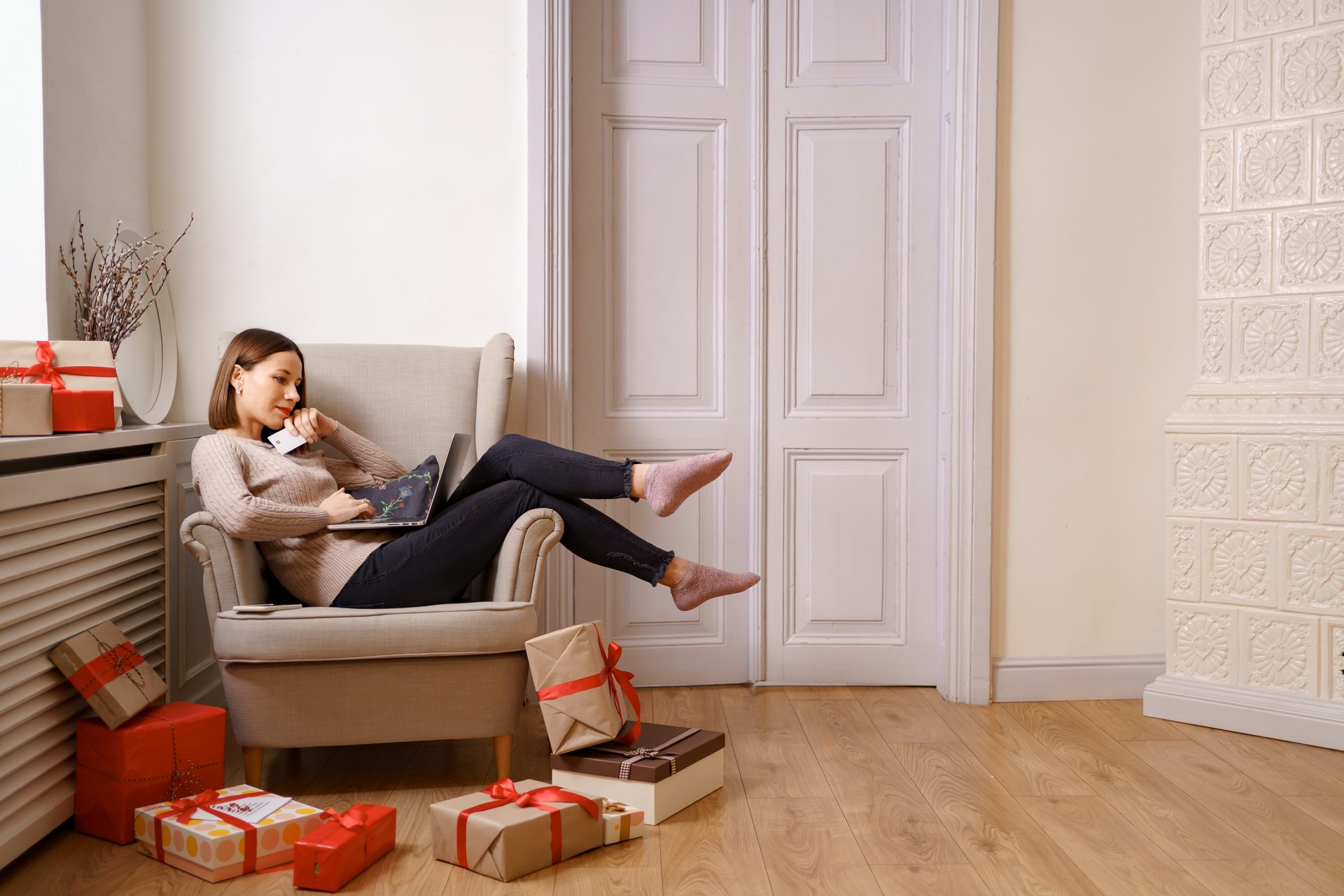 woman on chair with credit / debit card in hand and computer on lap. she is surrounded by presents.