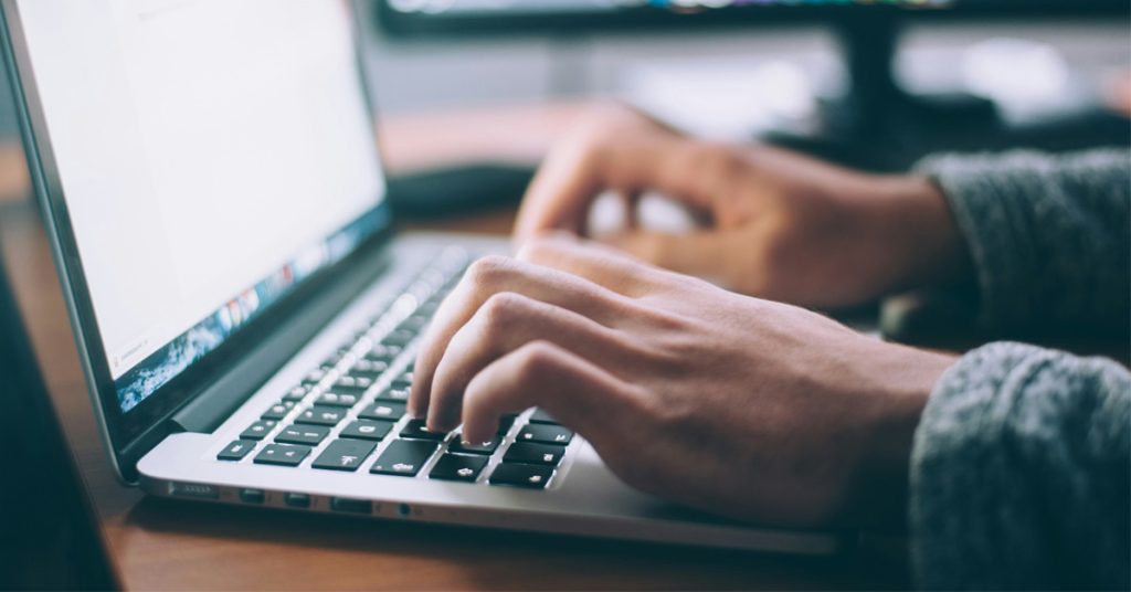 Two hands type on a keyboard as a person faces a blank document on the screen.