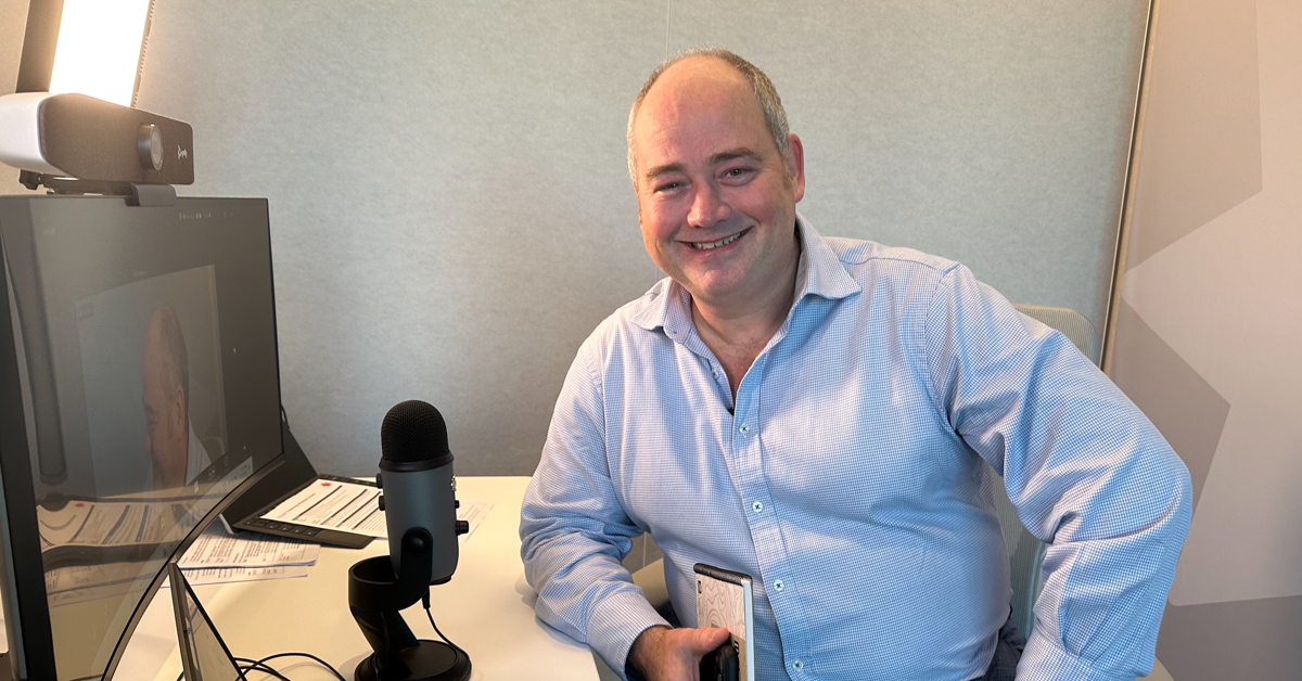 A man in a pale blue shirt sits at a desk, smiling and facing the camera. He holds a notebook and is surrounded by a microphone, light and screens.