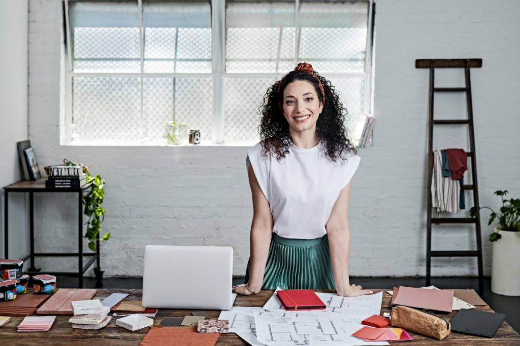 A woman stands at a desk with a laptop beside her. She is smiling and facing the camera in a white top.
