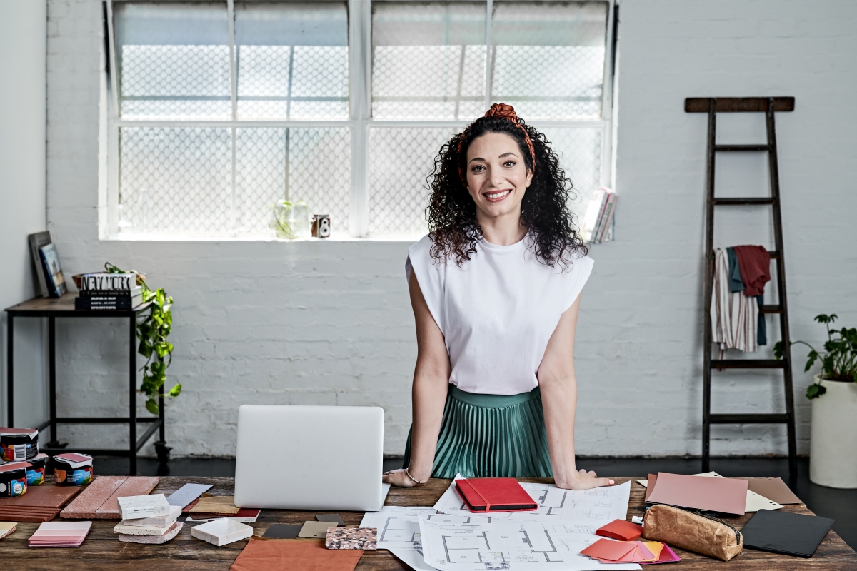 A woman stands at a desk with a laptop beside her. She is smiling and facing the camera in a white top.